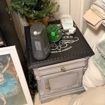 Sale Photo Thumbnail #278: Distressed grey painted nightstand with black top graphic, featuring small appliances including a portable space heater, desk fan, and white noise machine.