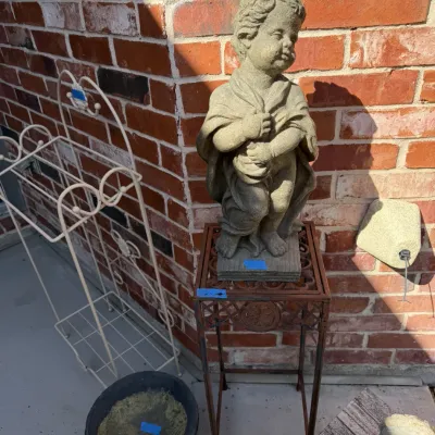 Sale Photo Thumbnail #395: Concrete garden statue of a young child wrapped in a blanket, displayed on a square metal plant stand alongside a white wire rack.
