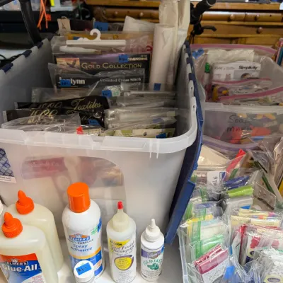 Sale Photo Thumbnail #359: Large assortment of crafting supplies, including clear plastic storage totes filled with cross-stitch kits, sewing patterns, fabrics, and various adhesives like Mod Podge and Elmer's glue.