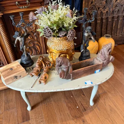 Sale Photo Thumbnail #131: Collection of decorative home decor displayed on a painted coffee table, including a gold Buddha head planter, cherub candelabras, carved wooden figures,