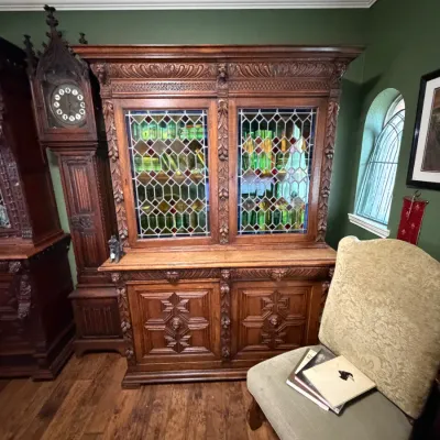 Sale Photo Thumbnail #4: Ornately carved antique oak hutch featuring leaded glass doors and intricate floral carvings, displayed alongside an antique grandfather clock.