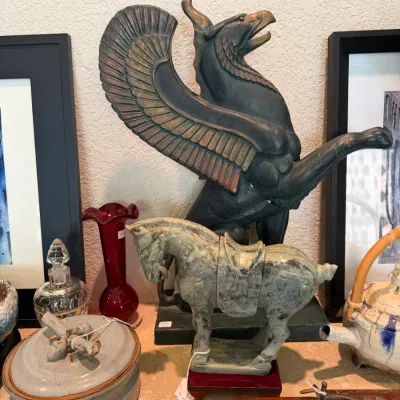 Sale Photo Thumbnail #156: Collection of decorative collectibles, including a large dark-finished griffin statue, a carved jade-style Tang horse figure, red art glass vase, and a studio pottery container.