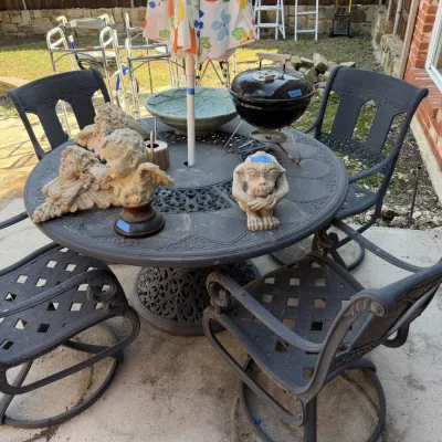 Sale Photo Thumbnail #408: Black wrought-iron outdoor dining table set with four swivel chairs, umbrella, small Weber charcoal grill, and stone garden statues.