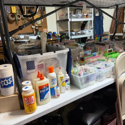 Sale Photo Thumbnail #356: Collection of crafting supplies including Mod Podge, Elmer's Glue-All, spray adhesives, and plastic storage tubs filled with assorted papercraft items.