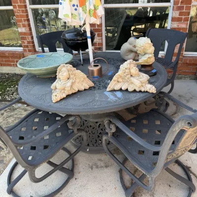 Sale Photo Thumbnail #389: Wrought-Iron patio dining set with a round lattice-top table, matching chairs, and a patio umbrella, displayed alongside stone angel statues and decorative garden items.