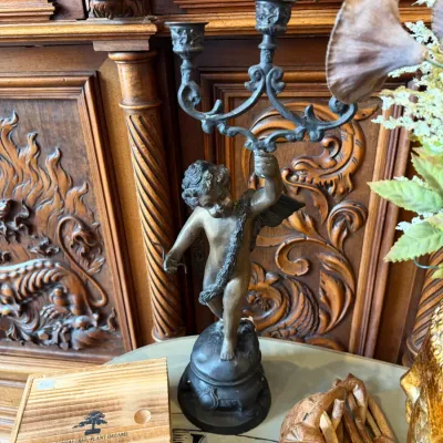 Sale Photo Thumbnail #132: Antique bronze cherub candelabra featuring a winged figure holding a two-arm scrolled candlestick (Pair) set against an ornate carved wooden cabinet 