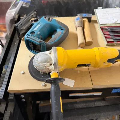 Sale Photo Thumbnail #326: Assortment of power tools, including a yellow DeWalt angle grinder, a teal Makita cordless jigsaw body, and wooden mallets on a workbench.