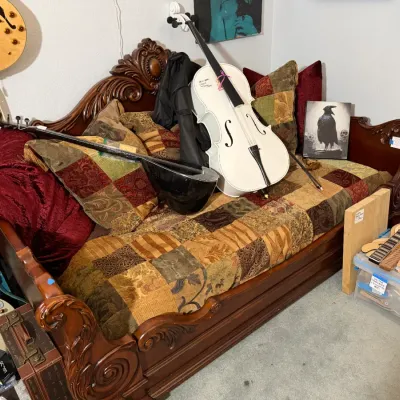 Sale Photo Thumbnail #116: Ornately carved wooden daybed with luxurious linens and pillows, featuring musical instruments displayed on top, including a white cello and a long-necked lute.