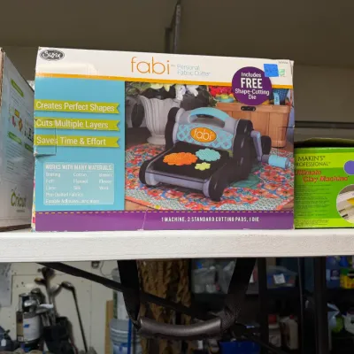 Sale Photo Thumbnail #366: Sizzix Fabi personal fabric cutter in its original box, displayed alongside a Makin's Professional Ultimate Clay Machine and a Cricut box on a shelf.
