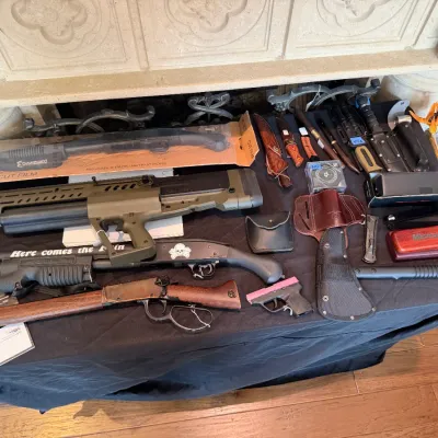Sale Photo Thumbnail #56: Collection of firearms, tactical gear, and edged weapons displayed on a table, including shotguns, a lever-action rifle, a compact handgun, fixed-blade knives, and a hatchet.