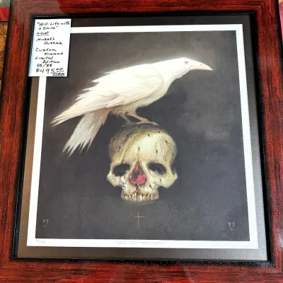 Sale Photo Thumbnail #173: Framed limited edition print, "Still-Life with a Smile" by Michael Hussar, numbered 55/88, featuring a white raven perched atop a skull in a reddish-brown wooden frame.