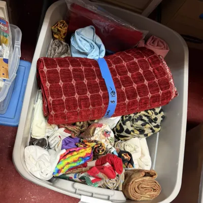 Sale Photo Thumbnail #369: Plastic storage tub filled with various fabric remnants, textiles, and sewing material, including a roll of red textured upholstery fabric.