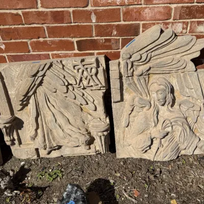 Sale Photo Thumbnail #400: Two-piece carved architectural relief panel depicting an angel with prominent wings, displayed outdoors against a brick wall.