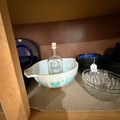 Sale Photo Thumbnail #260: Vintage Pyrex Butterprint Cinderella mixing bowl with a clear glass decanter, alongside clear pressed glass bowls and dark blue ceramic tableware inside a cabinet.