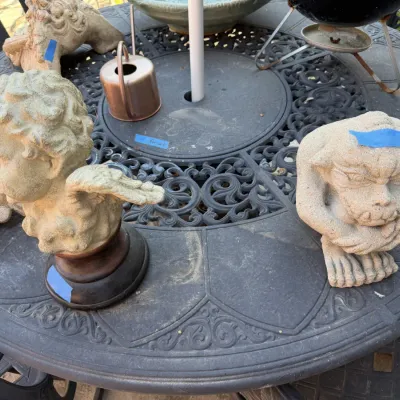Sale Photo Thumbnail #386: Assorted concrete garden decor, including cherub busts and a crouching gargoyle figure, displayed on a wrought-iron patio table.