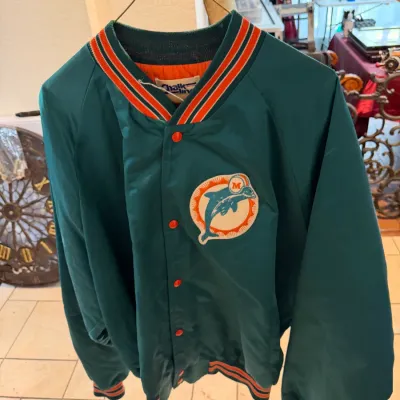 Sale Photo Thumbnail #32: Vintage 1980s Chalk Line Miami Dolphins satin bomber jacket featuring an embroidered logo patch, orange snap closure buttons, and striped knit trim.