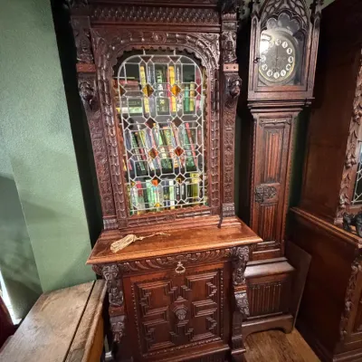 Sale Photo Thumbnail #3: 19th-century ornately carved Gothic Revival bookcase featuring a leaded glass door and intricate wood relief details, displayed alongside a matching tall case clock.