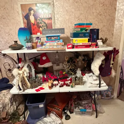 Sale Photo Thumbnail #272: Collection of estate sale items displayed on folding tables, featuring vintage board games, Art Deco style artwork, glassware, and holiday decorations.